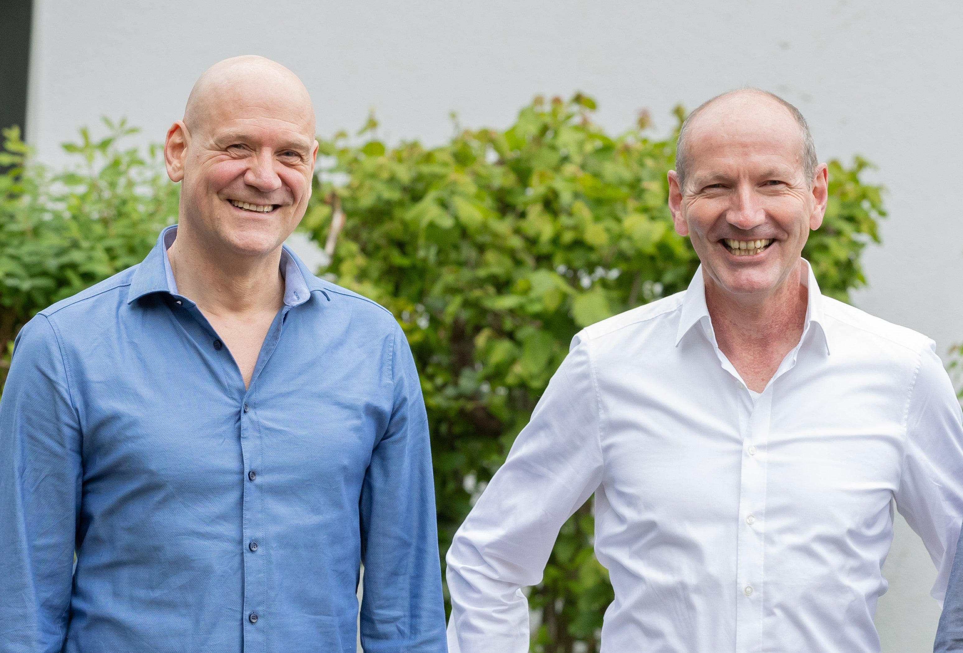 Two smiling men standing outdoors against a leafy background, one wearing a blue shirt and the other in a white shirt.