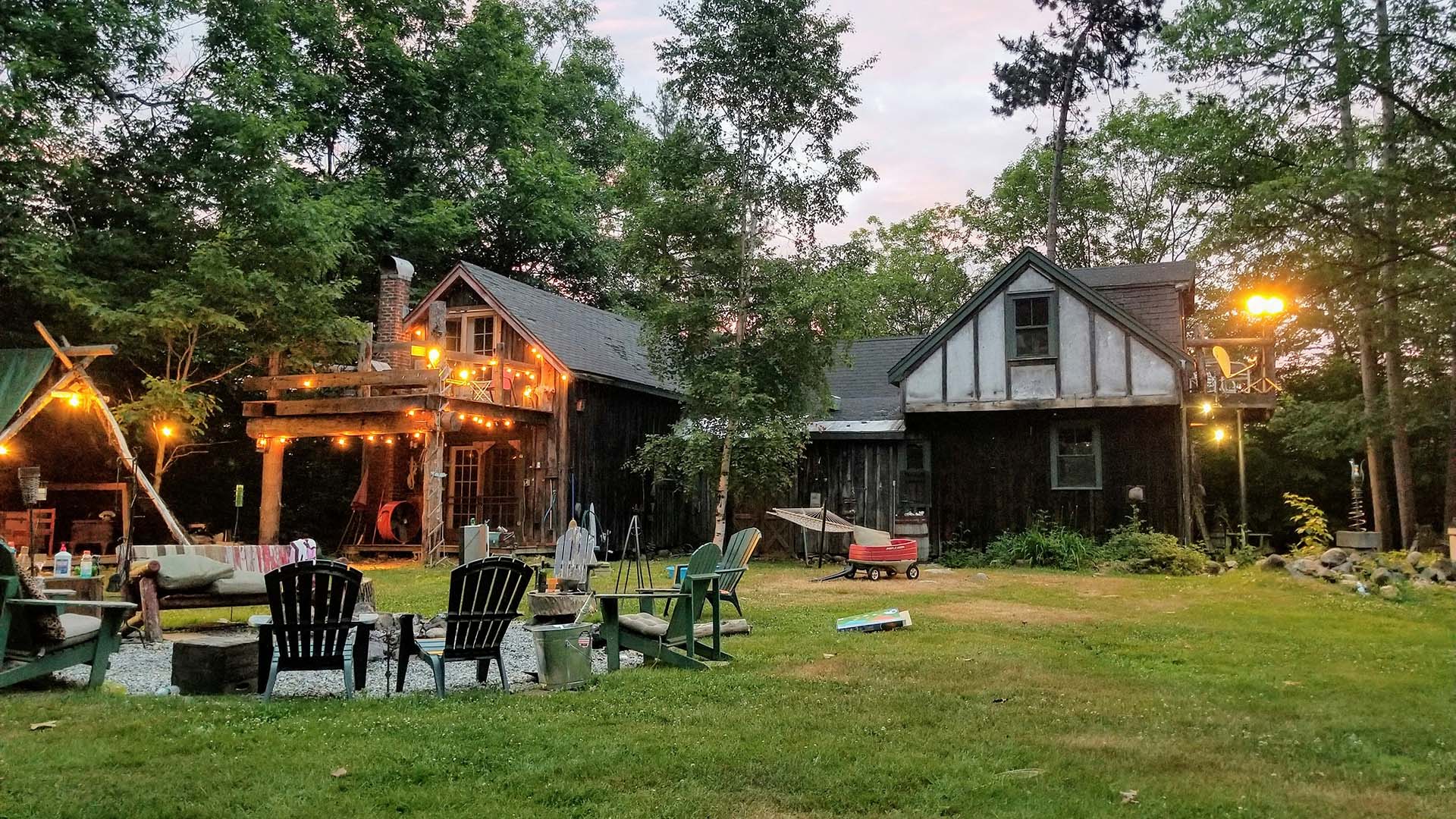 Rustic cabin in a wooded area with string lights, Adirondack chairs around a fire pit, and evening ambiance.