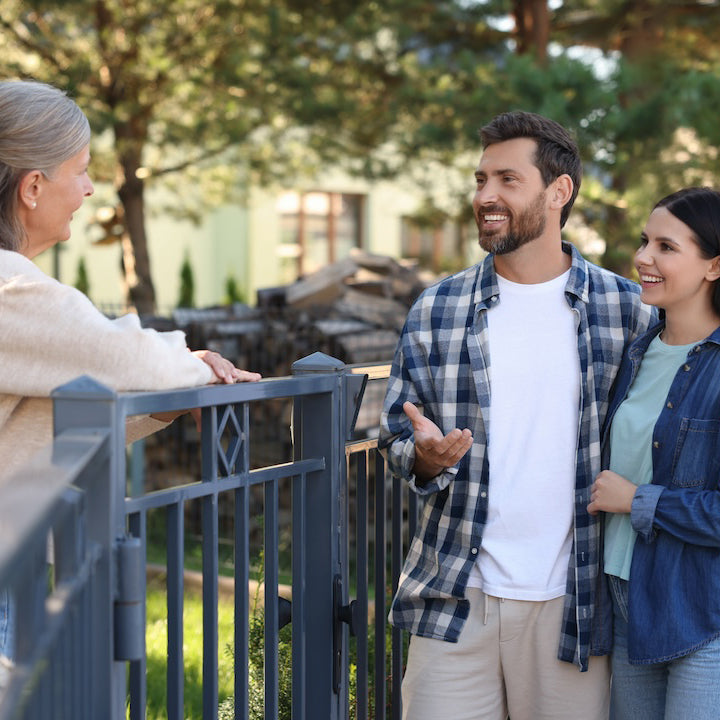 Friendly neighbors talking over a fence in a suburban setting with trees in the background.