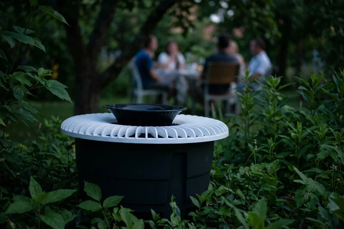 Dark outdoor mosquito trap with a ribbed light-colored top and black bowl among garden foliage.