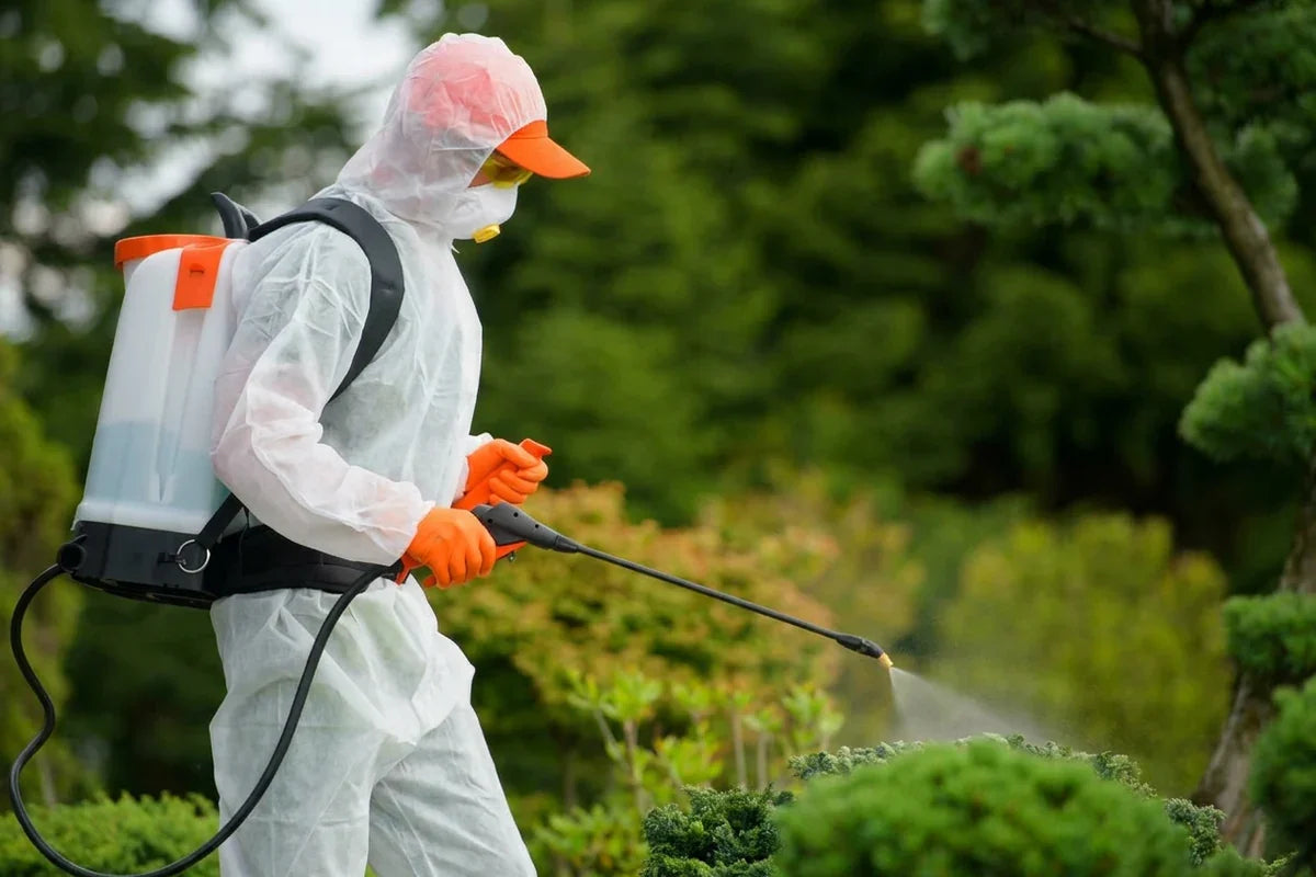 Person in protective clothing sprays vegetation with a backpack sprayer and handheld wand.